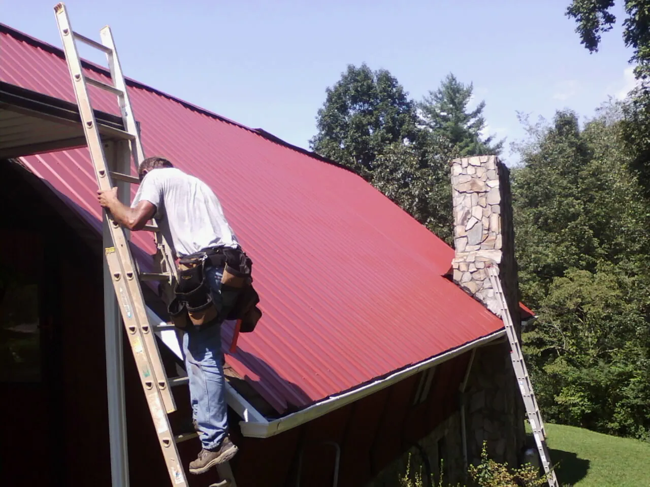 Roofer on a ladder beside a red metal roof and stone chimney — Soffit & Fascia Repair by Roofing Brothers in Artois